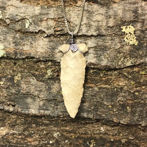 May include: A silver chain necklace with a light brown arrowhead pendant. The arrowhead is attached to the chain with a silver wire wrapped around the top of the arrowhead.