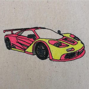 May include: An embroidered image of a sports car in yellow and pink on a beige fabric background.