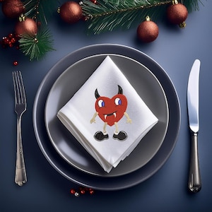 May include: A white napkin with a red heart-shaped devil embroidered on it. The napkin is folded and placed on a grey plate. The plate is on a blue surface.