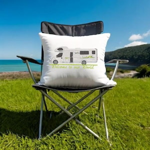 Könnte beinhalten: Weißes Kissen mit einer grünen und grauen Illustration eines Wohnmobils und dem Text "Welcome to our World". Das Kissen liegt auf einem Klappstuhl, vor dem Hintergrund von grünem Gras und blauem Himmel.