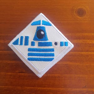 May include: Diamond-shaped embroidered patch with a robot design in blue and white. The patch has a white background with blue detailing, including stripes and a circular black eye. The edges are white.