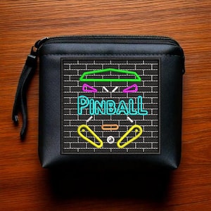 Può includere: Borsa nera con cerniera con un design ricamato in verde neon, blu, rosa e giallo di un flipper su un muro di mattoni. Il design dice "PINBALL" in lettere blu neon.