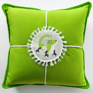 以下が含まれることがあります： 緑色の枕に、白い刺繍で囲まれた円の中に、黒い目の緑色のトカゲが描かれています。トカゲは白いアクセントが付いています。