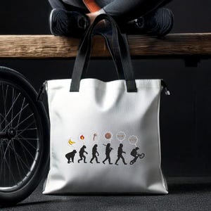 Può includere: Borsa tote in tela bianca con manici neri. La borsa presenta una grafica in bianco e nero dell'evoluzione dell'uomo, che termina con un uomo che guida una BMX. La grafica include bolle di pensiero con icone che rappresentano diverse fasi dell'evoluzione umana.