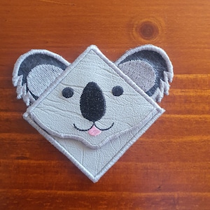 Könnte beinhalten: Ein grauer Koala-Bären-Aufnäher mit schwarzen Augen, einer schwarzen Nase und einer rosa Zunge. Der Koala hat graue Ohren mit einem strukturierten Design. Der Aufnäher ist rautenförmig und hat einen genähten Rand.
