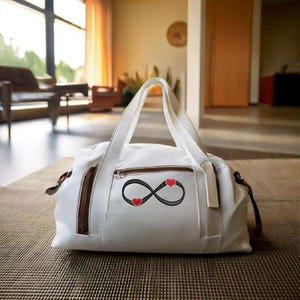 Könnte beinhalten: Weiße Canvas-Sporttasche mit braunen Lederelementen und Reißverschluss. Die Tasche hat ein gesticktes Unendlichkeitszeichen mit zwei roten Herzen.