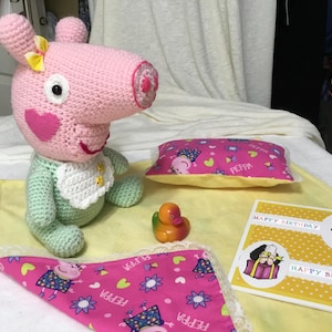 May include: A handmade pink crocheted pig doll with a green body, white bib, and yellow bow. Includes a pink pillow, blanket, rubber duck, and a "Happy Birthday" card. The items are on a yellow blanket.