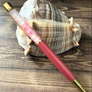 Può includere: Una penna decorativa con fusto rosa e dettagli dorati. La sezione superiore trasparente della penna è riempita di glitter rosa e bianchi. La penna è appoggiata su una conchiglia su uno sfondo di legno.