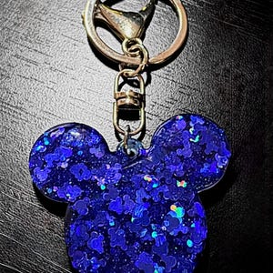 May include: A blue glitter keychain in the shape of Mickey Mouse's head. The keychain has a gold-colored clasp and ring. The glitter is a mix of blue and iridescent colors, creating a sparkling effect.