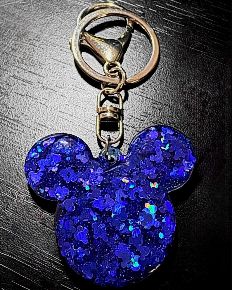 May include: A blue glitter keychain in the shape of Mickey Mouse's head. The keychain has a gold-colored clasp and ring. The glitter is a mix of blue and iridescent colors, creating a sparkling effect.