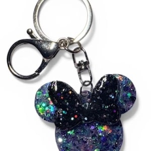 May include: A glittery Minnie Mouse-shaped keychain. The keychain features a black bow and a body filled with purple, blue, and green glitter. The keychain has a silver clasp and a keyring.