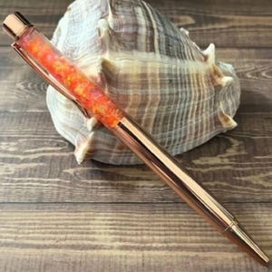 Puede incluir: Un bolígrafo de oro rosa con un barril transparente lleno de purpurina naranja y amarilla. El bolígrafo está sobre una superficie de madera junto a una gran concha. El bolígrafo tiene un diseño elegante y moderno.