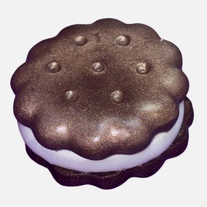 May include: A chocolate-colored, glittery, sandwich cookie with a white filling. The cookie has a scalloped edge and small, raised dots on the top. The overall effect is a whimsical, dessert-themed item.