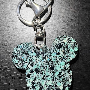 May include: A glittery Mickey Mouse-shaped keychain. The charm is a mix of black, teal, and silver glitter. The keychain has a silver clasp and ring for attaching to keys or a bag.