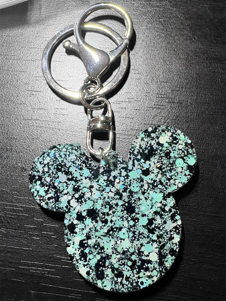 May include: A glittery Mickey Mouse-shaped keychain. The charm is a mix of black, teal, and silver glitter. The keychain has a silver clasp and ring for attaching to keys or a bag.