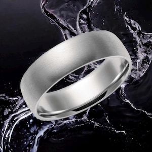 May include: A silver-toned metal ring with a brushed, matte exterior finish and a smooth, polished interior band. The ring features a simple, rounded profile and a solid construction, presented against a dark background with liquid splashes.