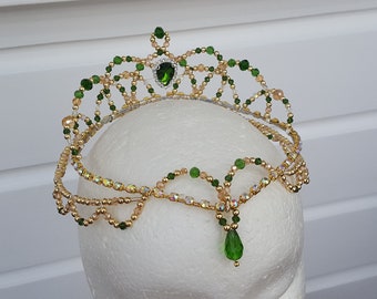 May include: A gold-toned beaded tiara with green and clear accents. The crown features a central green gem and a dangling teardrop-shaped gem. The design includes multiple strands of beads and is displayed on a white mannequin head.