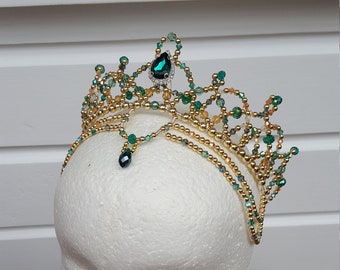 May include: A handcrafted gold-toned tiara with emerald green and clear crystal accents. The crown features a central emerald-colored jewel and dangling teardrop-shaped jewel. The tiara is displayed on a white foam head.