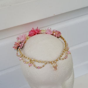 May include: A pink and gold floral crown with delicate beads and a wire base. The crown is adorned with pink fabric flowers and gold beads.