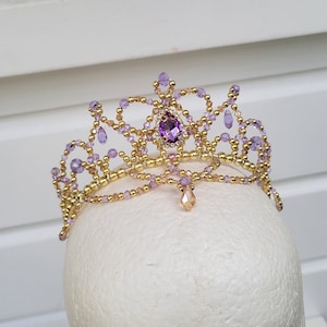 May include: A gold and purple beaded tiara with a large purple gemstone in the center. The tiara is on a white mannequin head.