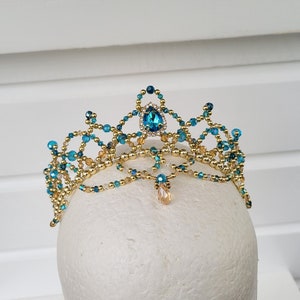 Puede incluir: Una tiara de cuentas doradas y azul verdoso con un gran cristal azul en forma de lágrima en el centro. La tiara está sobre una cabeza de maniquí blanca.