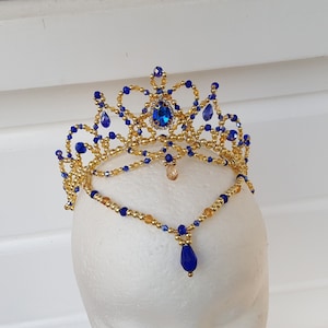 Puede incluir: Una corona de cuentas doradas y azules con un colgante en forma de lágrima. La corona está hecha con pequeñas cuentas doradas y cuentas de vidrio azules. El colgante es una cuenta de vidrio azul en forma de lágrima.