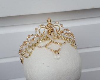 May include: A gold and white beaded tiara with a large crystal in the center. The tiara is on a white mannequin head.
