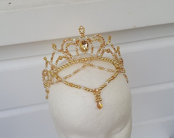 May include: A gold beaded crown with a teardrop-shaped crystal pendant. The crown is made of small gold beads and has a delicate design.