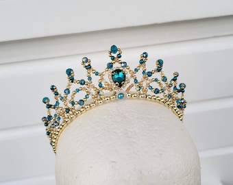 May include: A gold-colored beaded crown with teal and clear crystal accents. The crown features a large teal crystal at the center and is displayed on a white mannequin head. The design is intricate and elegant.