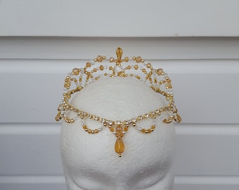May include: A gold and clear beaded crown with a teardrop pendant. The crown is on a white mannequin head.