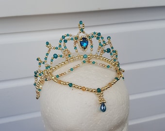 May include: A gold and teal beaded tiara with a large blue gemstone in the center. The tiara has a dangling blue gemstone drop.
