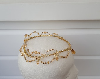 May include: A gold beaded crown with a delicate design. The crown is made with small gold and white beads and has a few dangling beads.