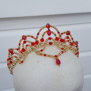 Puede incluir: Una tiara hecha a mano con una estructura de alambre dorado, adornada con cuentas rojas y doradas vibrantes. La corona presenta un diseño simétrico con colgantes de cristal rojo colgantes, perfecta para ocasiones especiales o disfraces.