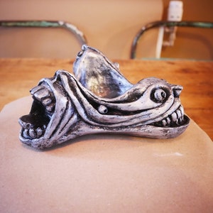 May include: A silver-colored, stylized sculpture of a snarling creature's head. The sculpture has sharp teeth and detailed facial features. It is placed on a piece of brown paper, with a wooden table and chairs in the background.