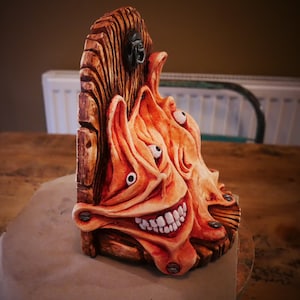 May include: A wooden wall shelf with a carved, grotesque face with multiple eyes and a wide, toothy grin. The shelf is painted in shades of brown and orange.
