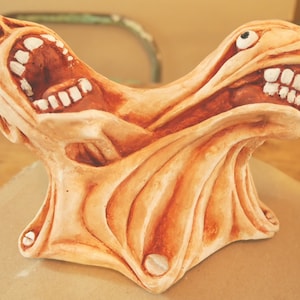May include: A ceramic sculpture of a grotesque, fleshy, human-like figure with two mouths open wide, revealing rows of teeth. The figure has a textured, wrinkled surface and appears to be melting or contorting.