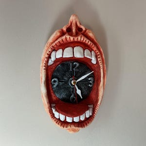 May include: A wall clock shaped like an open human mouth with a nose at the top. The interior of the mouth is red with white teeth and a tongue. A circular black clock face with white numbers 3, 6, 9, and 12 is set inside the open mouth.