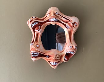 May include: A round mirror with a unique design featuring multiple faces with open mouths and teeth surrounding the reflective surface. The faces are painted in shades of peach and brown.