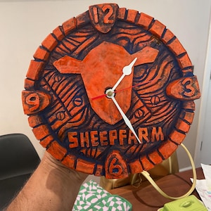 May include: A round wall clock with a textured orange and dark blue finish. The clock face features a central sheep head silhouette, the text SHEEPFARM, and raised numbers 12, 3, 6, and 9. It has white hour and minute hands and a red second hand.