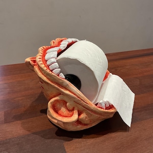 May include: A novelty toilet paper holder shaped like a grinning mouth with white teeth and red gums. The holder is made of a tan material and holds a roll of white toilet paper.