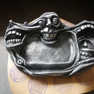May include: A silver-colored, grotesque face-shaped tray with multiple open mouths and exaggerated features. The tray is resting on a tan surface with purple stamps that read "Gaping Gobs".