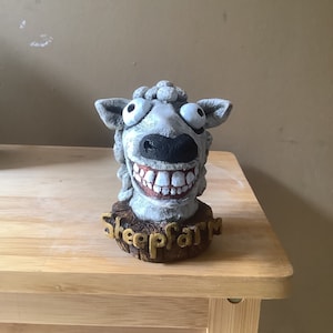以下が含まれることがあります： 大きな目と歯を見せる大きな笑顔など、誇張された特徴を持つ灰色の羊の頭の彫刻。彫刻は、金色の文字で「sheepfarm」という言葉が書かれた茶色の台座にあります。羊は、質感のあるウールのような外観をしています。
