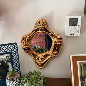 May include: A gold-colored mirror with a unique design featuring multiple faces with open mouths and teeth. The mirror is hanging on a white wall with a white thermostat next to it.