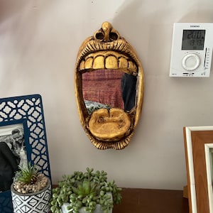 May include: A gold-painted, open-mouthed face mirror with a detailed design of teeth and lips. The mirror is mounted on a wall with a white background.