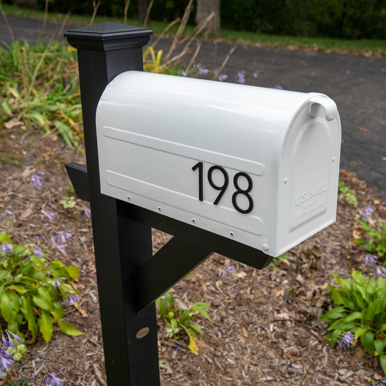 May include: A white metal mailbox mounted on a black post, featuring the black house numbers 198 on the side. The mailbox has a rounded top, a small handle on the front door, and the embossed text U.S. MAIL on the lower right corner of the door.