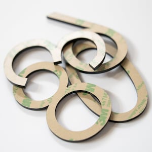 May include: A collection of black, circular, and curved adhesive-backed shapes. Each piece features a tan paper backing printed with green 3M 467MP adhesive branding. The items are stacked in an overlapping arrangement, showing a black edge.