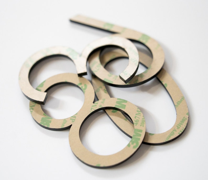 May include: A collection of black, circular, and curved adhesive-backed shapes. Each piece features a tan paper backing printed with green 3M 467MP adhesive branding. The items are stacked in an overlapping arrangement, showing a black edge.