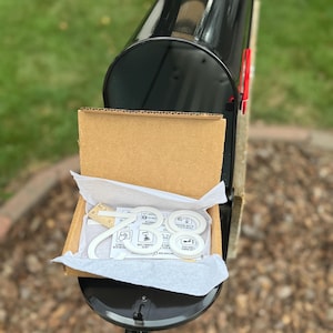 May include: A black metal mailbox with a cardboard shipping box resting inside. The box contains white house numbers, specifically the digits 2, 8, and 8, along with a small wooden ruler measuring approximately 2 inches in length for scale.