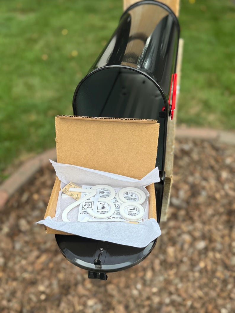 May include: A black metal mailbox with a cardboard shipping box resting inside. The box contains white house numbers, specifically the digits 2, 8, and 8, along with a small wooden ruler measuring approximately 2 inches in length for scale.
