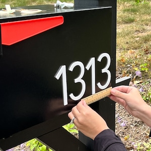 May include: A black mailbox featuring a bright red flag and the white house numbers 1313 attached to the side. A small wooden ruler is held against the bottom of the numbers to show their scale, measuring approximately 3 inches in height.